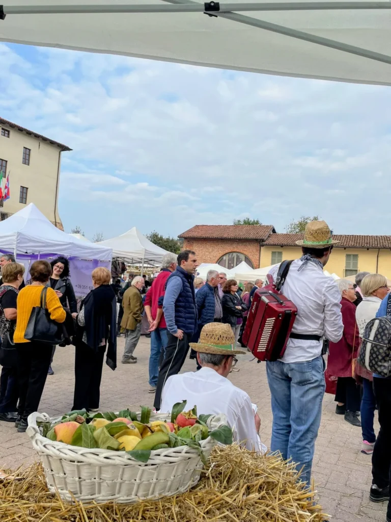 Fisarmonicista di spalle tra la gente, con un cesto di frutta autunnale in primo piano